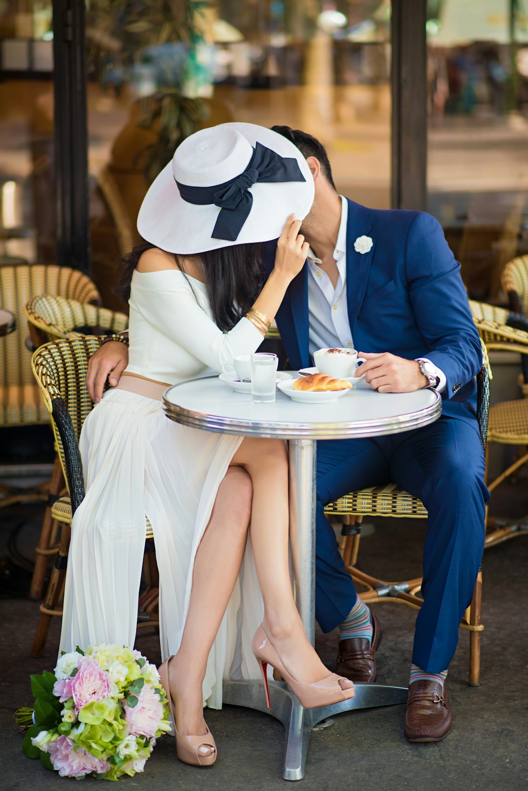 Stylish couple kissing behind a hat at a Parisian café, drinking cappuccino and eating croissants